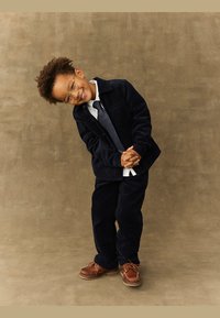 Marineblå cordfløyelsdress med to lommer, hvit skjorte og stripete slips. Brune sko med lisser. Barnet poserer lekent med et smil.
