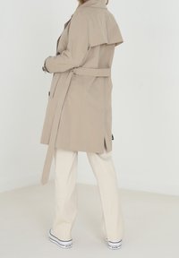 Beżowy trencz o podwójnym zapięciu, z klapą na plecach, wiązanym w pasie i bocznymi kieszeniami. Noszony na kremowe, szerokie spodnie i sneakersy.
