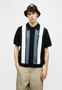 Νεαρός άνδρας που φοράει μαύρο καπέλο τύπου bucket, μαύρο πουκάμισο με κοντά μανίκια και κάθετες μπλε και λευκές ρίγες, και μπεζ παντελόνι, στέκεται ήρεμος.