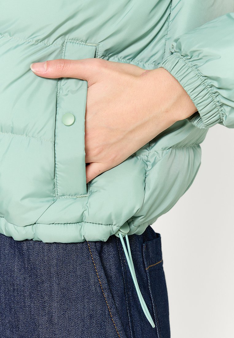Chaqueta acolchada color verde menta con una textura suave, que presenta una cintura elástica, un bolsillo lateral y un detalle de cordón ajustable.