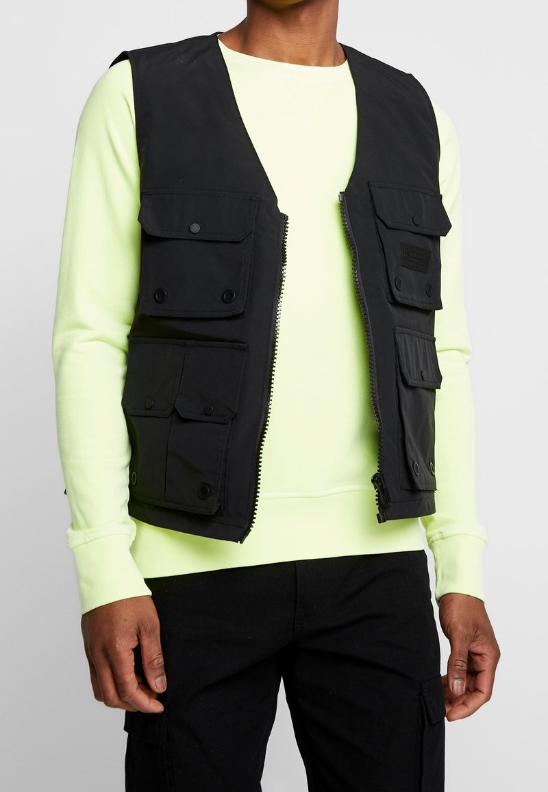 Svart väst med flera framfickor, dragkedjestängning och tryckknappar, bärs över en lysande neongrön långärmad skjorta.