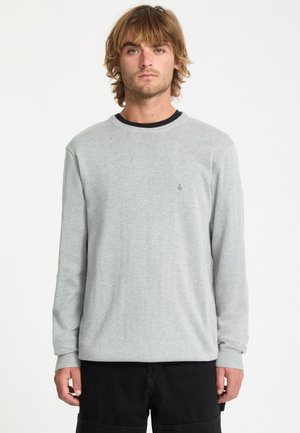 Jeune homme aux cheveux mi-longs, portant un pull léger gris clair à manches longues et un pantalon noir, debout devant un fond blanc uni.