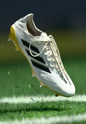 Weiße Fußballschuhe mit gelben Akzenten, schwarzem Streifen und einer strukturierten Oberfläche. Über Gras positioniert, mit fliegenden Rasenteilen.