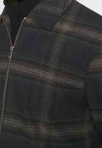 Tmavě zelená a hnědá kostkovaná bunda se zipem vpředu, nošená přes černý žebrovaný svetr.