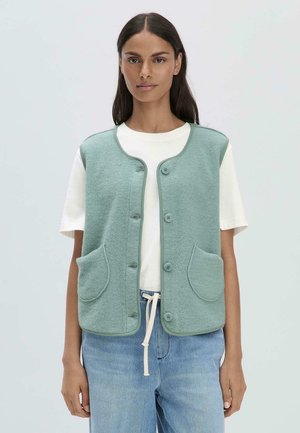 Hellgrüner Fleece-Weste mit rundem Ausschnitt, Fronttaschen und Knopfverschluss. Getragen über einem weißen T-Shirt und blauen Jeans. Glatte Textur.