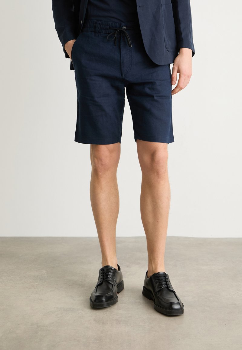 Pantaloni corti in lino blu navy con vestibilità rilassata e vita con coulisse. Abbinati a scarpe in pelle nere. Design slim-line, senza motivi o accenti visibili.