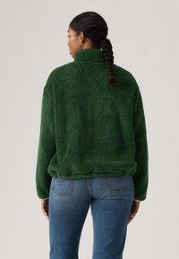 Γυναίκα με πλεξούδες φοράει πράσινο μπουφάν fleece και μπλε τζιν, εμφανιζόμενη από πίσω σε μονόχρωμο φόντο.