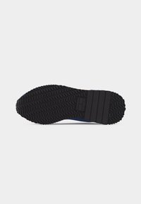 Semelle en caoutchouc noir de chaussure avec motif de grip texturé et éléments de design rectangulaires sur le talon, présentée sur fond gris clair.