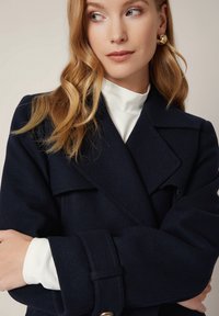 Granatowy wełniany płaszcz o podwójnym zapięciu, szerokich klapach i rękawach zapinanych na pasek. Nosi się go na białej bluzce z wysokim kołnierzem. Prosty, elegancki wygląd.