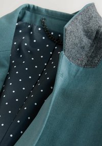 Ärmellose Jacke aus türkisfarbenem Außenstoff, grauem Wollkragen und navyblauer Innenausstattung mit weißen Punkten, Kettendetail im Kragen.