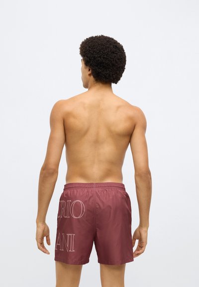 Bordó fürdőnadrág elasztikus derékrésszel és logófelirattal a háton, sima textúrával és térd alá érő kialakítással. Egyéb ruházat nincs rajta.