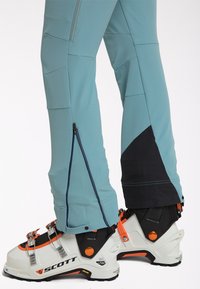 Pantalon de ski bleu clair avec renfort noir aux chevilles, porté au-dessus de chaussures de ski Scott blanches et orange avec boucles métalliques.