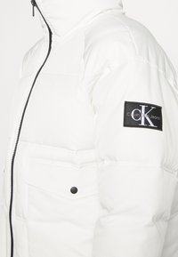 Giubbotto piumino bianco con zip nera, tasca a patta con bottone e patch del logo Calvin Klein Jeans nera sulla manica superiore.