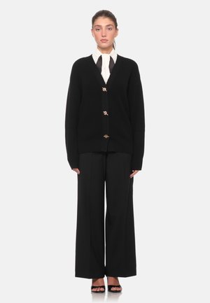 Donna che indossa un cardigan nero con bottoni dorati, camicetta bianca con colletto annodato, pantaloni larghi neri e sandali neri.