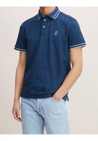 Laivastonsininen poolopaita, joka on valmistettu teksturoidusta kankaasta, ja jossa on kaulus ja kalvosimet, joissa valkoiset raidat, sekä rintalogo, jossa on vuosi 1962.