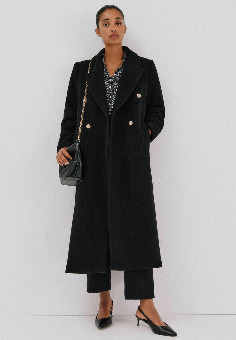 Musta villainen pitkä takki, jossa on leveät kaulukset, kaksiriviset napit, yhdistettynä kuvioituun paitaan ja mustiin housuihin. Asusteina on pieni laukku ja korot.