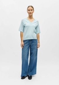 Femme debout, portant un haut bleu clair à col en V et manches courtes, un jean bleu à jambes larges, des chaussures noires, les cheveux attachés, sur un fond clair uni.