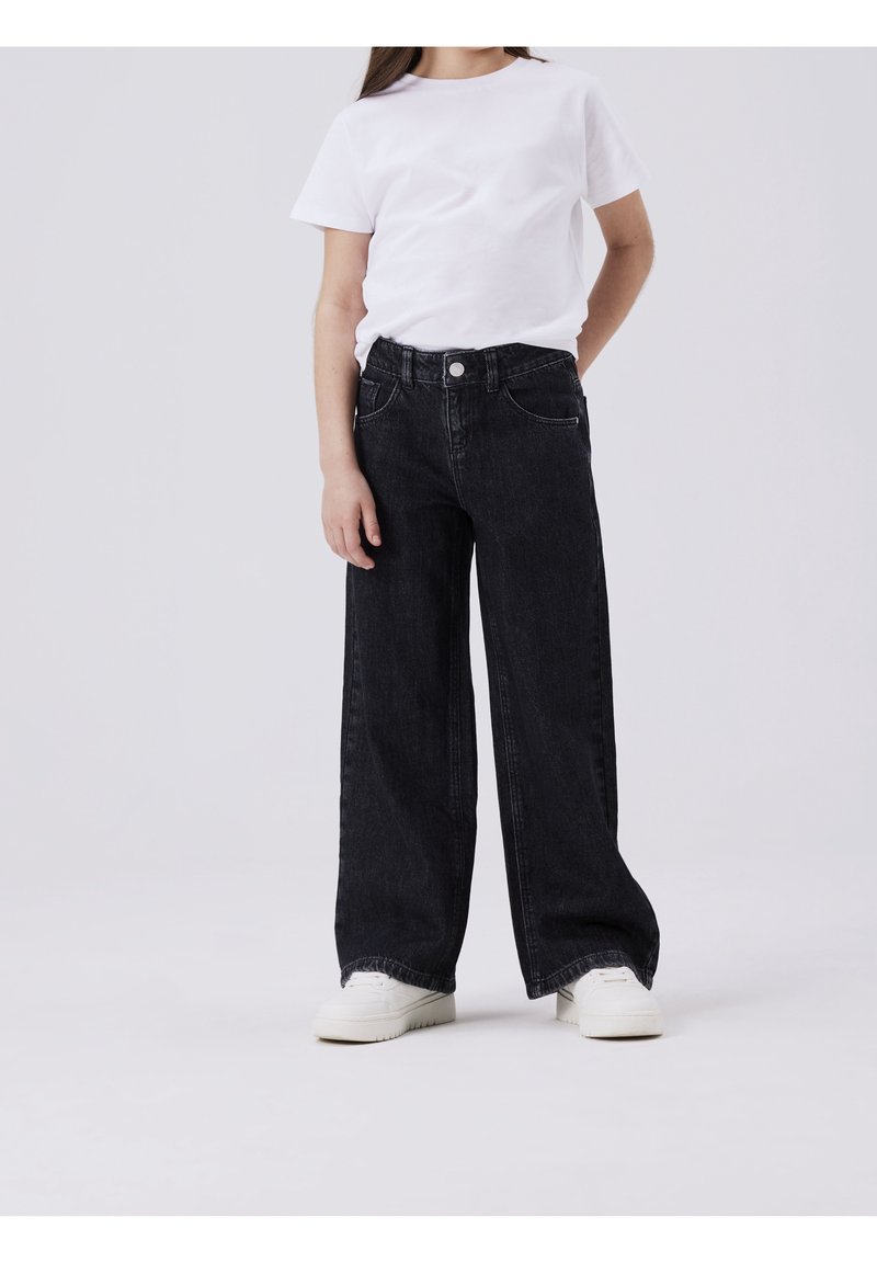 Enfant portant un t-shirt blanc uni à manches courtes, un jean noir à jambes larges et des baskets blanches, debout devant un fond clair uni.