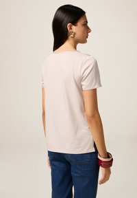 T-shirt rosa chiaro a maniche corte con scollatura rotonda, abbinato a jeans blu scuro, visto da dietro. Presenta un tessuto liscio e fessure laterali.