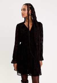 Svart blommönstrad klänning med V-ringning, genomskinliga långa ärmar, draperad midja och utsvängd kjol. Modell som bär stora tårformade örhängen.