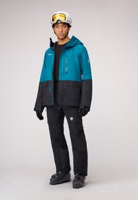 Skijacke in Türkis und Schwarz, mit einer Reißverschlusstasche auf der Brust, verstellbarem Kapuze und passenden schwarzen Skihosen mit Logos und strukturierten Akzenten.