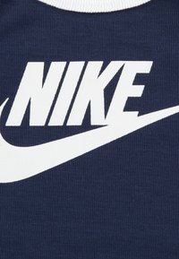 Tamsiai mėlyna medvilninė marškinėliai su dideliu baltu "NIKE" logotipu ir swoosh dizainu. Apykaklė yra baltos spalvos nėriniais, suteikiant kontrastą audiniui.