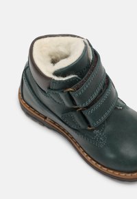 Botín de cuero verde hasta el tobillo con dos correas ajustables de velcro, un suave forro de lana y una suela marrón resistente. Textura lisa y diseño casual.