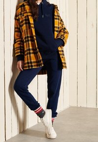 Chaqueta de franela a cuadros amarilla y negra sobre sudadera azul marino y joggers a juego, acompañada de zapatillas blancas y calcetines a rayas. Texturas suavizadas.