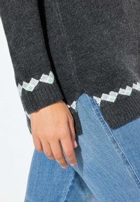 Maglione grigio a maglia con un motivo a rombi bianco e verde ai polsini, caratterizzato da uno spacco laterale, abbinato a jeans blu.