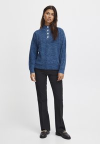 Pull en maille bleu à col monté et trois boutons clairs, associé à un pantalon noir à jambes droites et des chaussures noires avec un accent en chaîne.