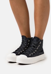 Para czarnych wysokich sneakersów z białymi plamkami, białymi gumowymi podeszwami i czarnymi sznurowadłami noszonych na gołych nogach na białym tle.