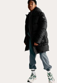 Zwarte gewatteerde jas met een capuchon en logo-opdruk, gedragen over een blauwe sweatshirt en grijze trainingsbroek. Witte sneakers met groene accenten.