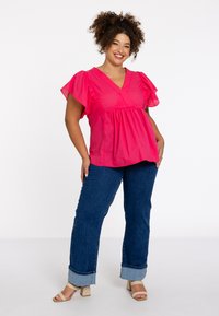 Rosa Baumwollbluse mit kurzen Rüschensäumen und V-Ausschnitt, kombiniert mit dunkelblauen Jeans und beigen Blockabsatzsandalen.