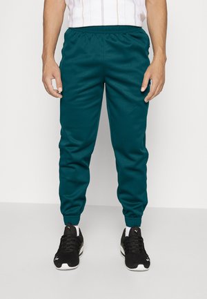 Pantalon de jogging fuselé couleur bleu sarcelle avec taille et poignets élastiques, fabriqué dans un tissu lisse. Associé à des chaussures de sport noires avec des accents blancs.