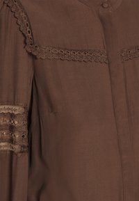 Tissu marron avec bordure en dentelle le long du yoke et de la manche, avec un bouton recouvert et des panneaux en dentelle texturés et semi-transparents sur la manche.