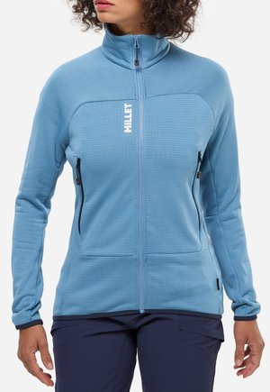 Frau trägt eine hellblaue Fleecejacke mit Reißverschluss und weißem "MILLET"-Schriftzug sowie dunkelblaue Hose, steht vor weißem Hintergrund.
