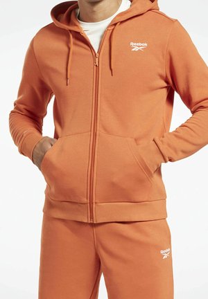 Mand iført orange Reebok lynlåshættetrøje og matchende joggingbukser, hænderne i lommerne, hvid skjorte synlig under hættetrøjen.