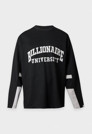 Jeune homme debout les mains dans les poches, portant un sweat-shirt noir « Billionaire University », un jean bleu foncé et des chaussures noires et blanches.