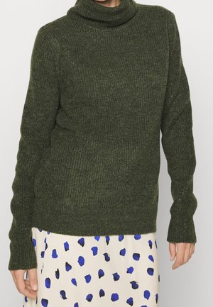 Vrouw draagt een donkergroene geribbelde coltrui en een witte rok met blauwe en zwarte abstracte vlekken.