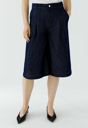 Frau trägt dunkle Denim-Weitbein-Shorts, weißes Top und schwarze spitze Slingback-Schuhe, steht mit Händen in den Taschen.