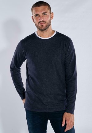 Dunkelblauses Langarm-T-Shirt mit einem weißen Rundhalsausschnitt, strukturiertem Stoff und lässiger Passform, kombiniert mit blauen Jeans.