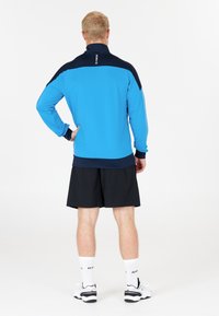 Ljusblå zip-up jacka med marinblå detaljer, långa ärmar och hög krage, tillsammans med svarta shorts och vita sportskor.