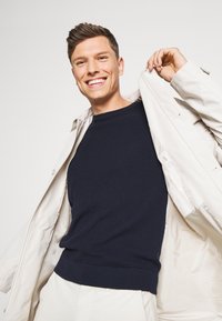 Mørkeblå strukturert strikkegenser med ribbet kant, brukt under en lys beige jakke. Modellen smiler mens hun holder jakken åpen.