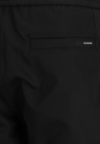 Pantaloni neri con una texture liscia, caratterizzati da una tasca posteriore e una piccola etichetta "CHASIN'" per un tocco di branding. Design semplice e funzionale.