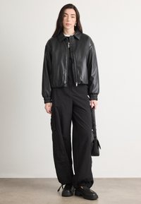 Giacca bomber in pelle nera con zip e polsini elasticizzati. Pantaloni cargo neri con tasche laterali, abbinati a scarpe nere chunky e una borsa piccola.