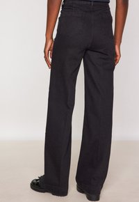 Persona che indossa pantaloni a vita alta neri con gamba larga e scarpe chunky nere, in piedi contro uno sfondo chiaro e uniforme.