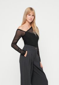 Top lungo a maniche nere in rete con scollo asimmetrico e corpo nero opaco, abbinato a pantaloni grigi plissettati a vita alta.