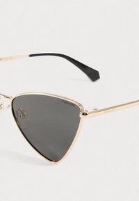 Γυαλιά ηλίου με χρυσό μεταλλικό σκελετό και μαύρους φακούς, τριγωνικού σχήματος, με κομψές μαύρες ακίδες. Περιλαμβάνει το λογότυπο "Polaroid" στον φακό.