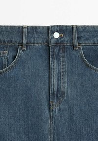 Vue de face de la taille d’un jean en denim bleu avec fermeture par bouton, passants de ceinture et deux poches avant visibles.