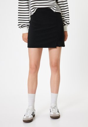 Schwarzer Minirock mit einem seitlichen Schlitz, aus glattem Stoff. Kombiniert mit einem gestreiften Oberteil, weißen Socken und Sneakers im Retro-Stil.